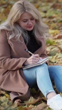 Dikey video. Mektup yazıyorum. Romantik kadın. Sonbahar ilhamı. Yaprakların üzerinde oturan gülümseyen bayan parktaki deftere notlar yazıyor..