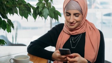 Kafe gezintisi. Akıllı telefon eğlencesi. Tesettürlü neşeli bir kadın telefon uygulamasında sohbet ediyor. Rahat bir kahve evinde dinleniyor..
