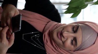 Dikey video. Cafe mobile. Akıllı telefon eğlencesi. Tesettürlü neşeli bir kadın telefon uygulamasında sohbet ediyor. Rahat bir kahve evinde dinleniyor..