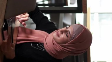 Dikey video. İnternetten alışveriş. İnternet ödemesi. Başörtüsü içinde güzel bir kadın dizüstü bilgisayarında kredi kartıyla sipariş alıyor. Kafedeki dijital satın alma bankacılığı hizmeti..