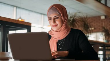 Uzaktan kumanda. Serbest çalışan biri. Gülümseyen, tesettürlü, tesettürlü, dizüstü bilgisayarda yazan, kahve dükkanındaki interneti karıştıran iş kadını..