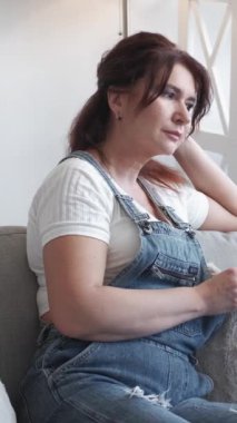 Dikey video. Romantik bir ruh hali. Hayalperest kadın. Evde rahatla. Gülümseyen orta yaşlı kadın, gündelik görünümlü, şezlongda oturmuş, aydınlık odada kahve içiyor..