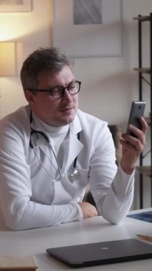 Dikey video. Tıbbi uzaktan tavsiye. Sağlık danışmanlığı. Çevrimiçi toplantı. Cep telefonu konferansında konuşan sağlık uzmanı kliniğin içindeki masada oturuyor..