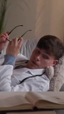 Dikey video. Sıkı çalışma. Yorgun doktor. Yorgun hastane uzmanı, klinikteki iş yerinde dizüstü bilgisayarla koltukta uyuyakalıyor..