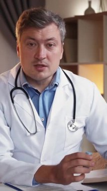 Dikey video. Doktor tavsiyesi. İlaç danışmanlığı. Profesyonel yardım. Tıbbi üniformalı olumlu bir adam kliniğin iç kısımlarında oturan ilaçların dozunu açıklıyor..