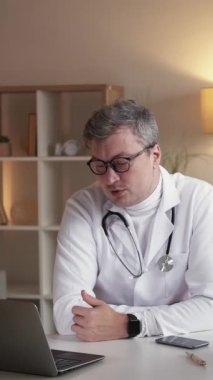 Dikey video. Sanal danışma. TeleHealth çağrısı. Dost canlısı doktor bilgisayarda konuşuyor. İnternetteki randevusu modern iç mekandaki çalışma masasında oturuyor..