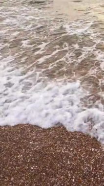 Dikey video. Sahil şeridi geçmişi. Okyanus dalgaları. Yaz tatili. Beyaz köpüklü berrak deniz suyu kahverengi kumsal deniz manzarasını süpürüyor..