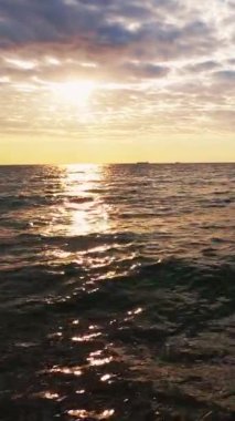 Dikey video. Deniz kenarı geçmişi. Günbatımı silüeti. Doğa manzarası güzelliği. Dalgalı su yüzeyi, dalga dalga güneş ışığını bulutlara yansıtıyor..