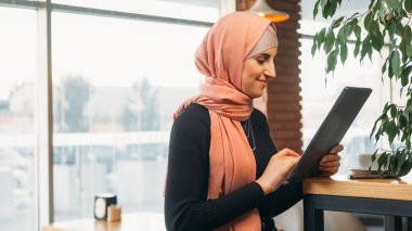 Cafe freelance. İnternet kullanıcısı. Tesettürlü neşeli bir kadın, dijital tablet ağında sohbet ediyor, rahat kahve dükkanında sosyal ağlara takılıyor, fotokopi çekiyor..