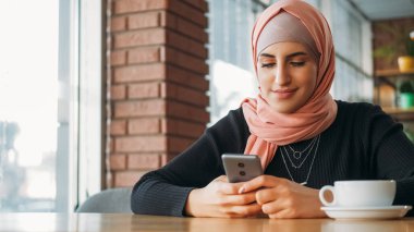 Cep telefonu kullanıcısı. Kahve keyfi. tesettürlü gülümseyen kadın akıllı telefondan sohbet mesajı yazıyor kafenin boş alanındaki internet uygulamasında geziniyor.