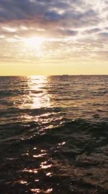 Dikey video. Deniz kenarı geçmişi. Günbatımı silüeti. Doğa manzarası güzelliği. Dalgalı su yüzeyi, dalga dalga güneş ışığını bulutlara yansıtıyor..