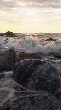 Dikey video. Sahil şeridi arka planı. Gündoğumu silüeti. Deniz kenarı doğası. Köpüklü su dalgaları bulut ufkunda kahverengi gri kayalara çarpıyor..