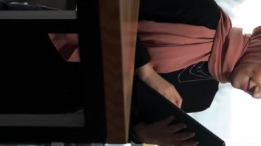 Dikey video. Uzaktan kumanda. İnternet taraması. Kafede oturmuş dijital tablet kullanarak sosyal ağları kontrol eden tesettürlü çekici bir kadın..