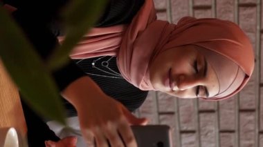 Dikey video. Mobil blog. İnternet araması. Mutlu tesettürlü kadın rahat kahve dükkanında sosyal ağda cep telefonuyla internet sohbeti yapıyor..