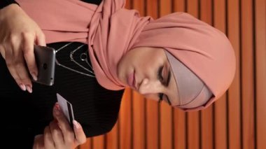 Dikey video. Cep telefonu ödemesi. İnternet alışverişi. Kafe 'de kredi kartı para transferi yapan tesettürlü çekici bir kadın telefon bankacılığı uygulamasından internetten sipariş veriyor..