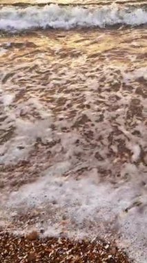 Dikey video. Deniz dalgası. Sahil şeridi arka planı. Deniz doğası manzarası. Beyaz köpüklü temiz su kahverengi kumlu okyanus kıyısında süzülüyor..