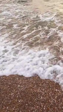 Dikey video. Sahil şeridi geçmişi. Okyanus dalgaları. Yaz tatili. Beyaz köpüklü berrak deniz suyu kahverengi kumsal deniz manzarasını süpürüyor..