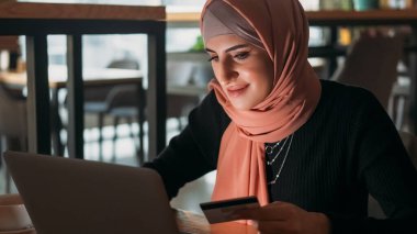 Çevrimiçi ödeme. İnternet alımı. Tesettürlü, tesettürlü bir kadın, kahve dükkanından kredi kartı e-ticaret para transferiyle laptoptan sipariş veriyor..