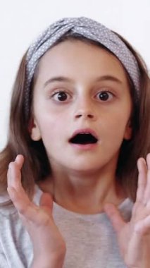 Dikey video. Şok olmuş çocuk. Şaşkın çocuk. Korkmuş korkmuş küçük bir kız, beyaz arka planda izole edilmiş ellerle ağzını kapatıyor..