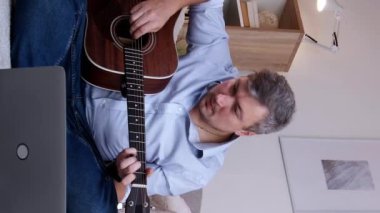 Dikey video. Gitar öğrencisi. Sanal eğitim. İnternet müzik dersi. Melodi çalan mutlu, ilham verici adam evde dizüstü bilgisayar konferansında oturmuş başarının tadını çıkarıyor..