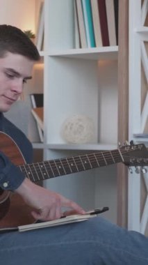 Dikey video. Şarkı yazmak. Müzik bestecisi. Akustik gitar. Yetenekli genç adam, müzik aleti ile evde oturma odasındaki günlükte melodi yaratıyor..