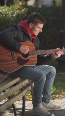 Dikey video. Sokak gitaristi. Müzisyen yaşam tarzı. Gençlik kültürü. Akustik enstrüman çalan yetenekli bir adam güneşli parktaki bankta eğleniyor..