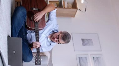 Dikey video. Gitar eğitimi. İnternet dersi. Uzaktan müzik kursu. Odaklanmış adam, akustik enstrüman eğitimi alıyor. Dizüstü bilgisayarlı, modern ev yapımı, yer fotokopi alanı..