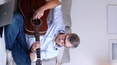 Dikey video. Gitar dersi. Mesafe dersi. Uzaktan eğitim. Akustik enstrüman çalan zeki bir adam ev içi fotokopi alanında dizüstü bilgisayarla yerde oturuyor..