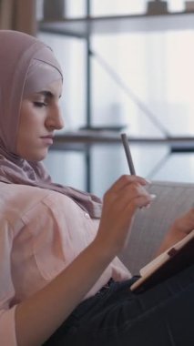 Dikey video. Günlük boş zaman. Hobi yazıyorum. Hafta sonu evde. Düşünceli, ilham verici kadın öğrenci rahat koltukta günlük planlaması yapıyor..