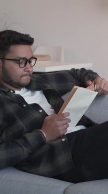 Dikey video. Kitap okumak rahatlatıcı. Bilgi ve rahatlık. Ev istirahatı. Gözlüklü barışçıl öğrenci oturma odasındaki rahat kanepede hafta sonunun tadını çıkarıyor..