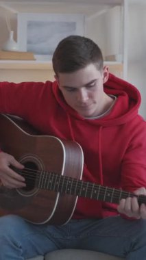 Dikey video. Müzik alıştırması. Profesyonel gitarist. Yetenekli bir sanatçı. Evdeki kanepede akustik enstrüman çalan ilham verici genç bir adam..