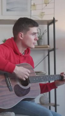Dikey video. Erkek gitarist. Müzikal eğlence. Melodi çalışması. Evde oturma odasındaki kanepede akustik enstrümanla şarkı söyleyen yetenekli hayalperest adam..