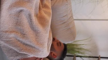 Dikey video. Evde şekerleme. Pazar dinlenmesi. Haftasonu rahatlama. Tembel bir gün. Modern oturma odasının iç kısımlarında battaniyenin altında rahat koltukta uyuyan yorgun adam..