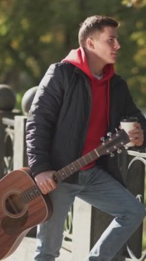 Dikey video. Açık hava yürüyüşü. Kahve molası. Şehir yaşam tarzı. Gitarlı hayalperest bir müzisyen Sunny Park caddesinde latte içerek duvar manzarasının tadını çıkarıyor..
