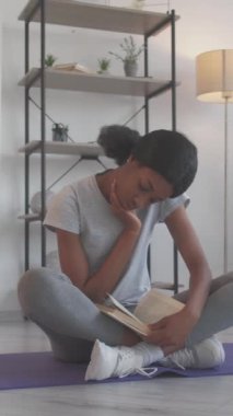 Dikey video. Fitness kitabı. Boş vakitleri değerlendiriyorum. Refah yaşam tarzı. Yoga dersine odaklanmış kadın oturma odasındaki pilates paspası üzerine kitap okuyor..