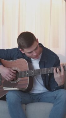 Dikey video. Gitar alıştırması. Müzik öğrenme. İlham eksikliği. Motive olmuş adam evdeki kanepede akustik enstrümanla melodi akorlarıyla boğuşuyor..