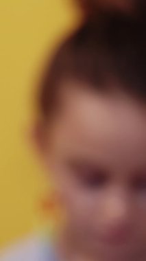 Dikey video. Çocuk yüzü. Ciddi çocuk. Mutsuz bakışlı, sahte parlak çilli, renkli küpeli küçük bir kız. Sarı arka plan 2 'de bulanık görünüyor..