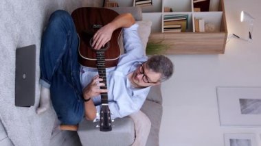 Dikey video. Evde eğitim. Müzisyen öğrenci. İnternetten ders. Akıllı konsantre adam, akustik gitar akorları üzerinde çalışıyor. Oturma odasının zemininde oturmuş, fotokopi çekiyor..
