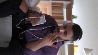 Dikey video. Sanal sohbet. Çevrimiçi çağrı. Dijital iletişim. Kulaklık takan kendine güvenen bir adam modern iç mekandaki rahat koltukta telefonla konuşuyor..