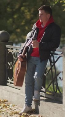 Dikey video. Açık havada kahve. Paket servis içkisi. Sokak müzisyeni. Gitarlı hayalperest genç adam güneşli bir sonbahar sabahı parkında sıcak içeceğinin tadını çıkarıyor..