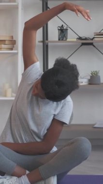 Dikey video. Spora yeni başlamış. Sabah sporu. Aktif kıyafetli, yoga minderinde sırt kaslarını esneten, oturma odasında eğlenen motive olmuş genç bir kadın..