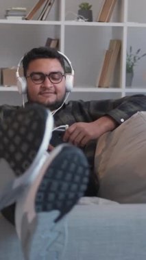 Dikey video. Müzik zevki. Ses aygıtı. Çalma listesi eğlencesi. Ev tatili. Mutlu gülümseyen adam telefonda en sevdiği şarkıyı dinliyor koltukta, rahat oturma odasında..