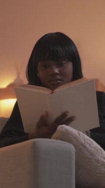 Dikey video. Evde öğrenim. Bilgi bilgeliği. Zeki düşünceli kadın modern oturma odasındaki koltukta açık kitap okumaktan zevk alıyor..