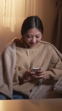 Dikey video. Müzik uygulaması. Hafta sonu çalma listesi. Samimi bir ortam. Rahatlamış mutlu kadın cep telefonu ev içini kullanarak kulaklıklarda mp3 sesi dinliyor..