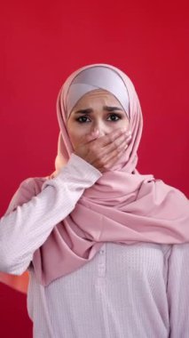 Dikey video. Omg şoku. Üzüntü. Acıma duygusu. Üzüntülü, tesettürlü, elleri kırmızı fotokopi arkaplanında izole edilmiş ağzı olan üzgün bir kadın..