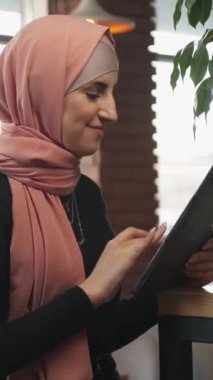 Dikey video. Cafe freelance. İnternet kullanıcısı. Neşeli kadın tesettürlü dijital tablet ağında sohbet ediyor rahat kahve dükkanında sosyal ağlara takılıyor.