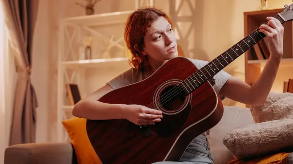 stock image Music practice. Guitar hobby. Inspired musician girl enjoying playing chords on string acoustic instrument on couch at home living room with free space.