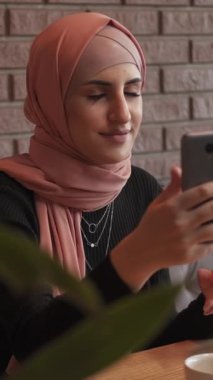 Dikey video. Mobil blog. İnternet araması. Mutlu tesettürlü kadın rahat kahve dükkanında sosyal ağda cep telefonuyla internet sohbeti yapıyor..