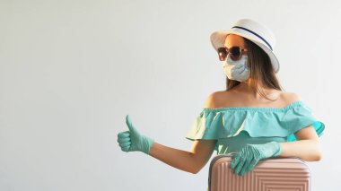 Salgın hastalık gezisi. Covid gibi. Tıbbi maske takan bir kız. Eldivenli, valizli, baş parmağıyla onaylı beyaz arka planda izole edilmiş boş bir yere bakıyor..