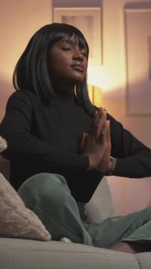 Dikey video. Evde yoga. Hafta sonu meditasyonu. Ruh uyumu. Lotus namaste pozisyonunda gözleri kapalı sakin bir kadın oturma odasının iç kısmında kanepede..